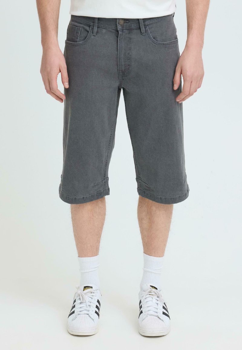 Homme portant un short en denim gris foncé arrivant aux genoux, des chaussettes blanches et des baskets Adidas blanches avec des rayures noires, debout devant un fond uni.