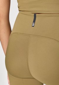 Olivno zelene športne legice z gladko teksturo, visokim pasom in majhno črno blagovno oznako na hrbtni strani. Vidne so šive.