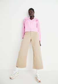 Blusa a maniche lunghe rosa chiaro con scollatura arricciata, abbinata a pantaloni beige larghi e sneakers multicolori, su uno sfondo bianco.