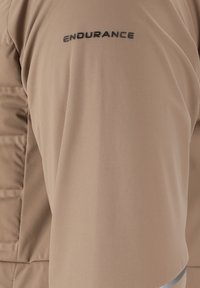 Beige Sportoberteil mit glatter Textur. Mit dem "ENDURANCE"-Logo in Schwarz auf der Schulter. Enthält einen grauen Akzent am Saum.