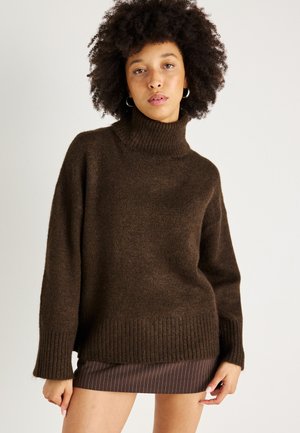 Pull encolure montant tricoté marron avec des poignets et un ourlet côtelés, associé à une jupe marron à rayures. Texture douce et coupe décontractée.