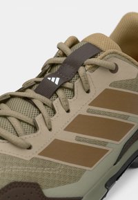 Αθλητικό παπούτσι σε αποχρώσεις ταμπά και ελιάς με αναπνεύσιμο επάνω μέρος από δίχτυ, υφασμένους λεπτομερείς τόνους, καφέ ρίγες και καφέ σύστημα κορδονιών.