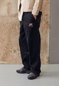 Svarta kordfläskbyxor med en broderad orange Nike-logotyp, kombinerat med grå sportskor, mot en texturerad betongbakgrund.