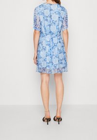 Robe florale bleue à manches courtes et taille nouée. Tissu en dentelle transparente et ourlet évasé. Portée avec des chaussures à talons hauts noires.