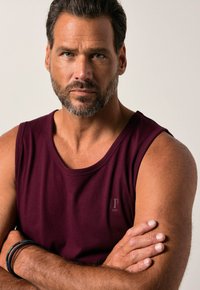 Bordowy top na ramiączkach z okrągłym dekoltem, z małym logo po lewej stronie klatki piersiowej. Tkanina wydaje się miękka i gładka w dotyku.