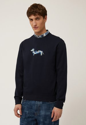 Felpa navy con collo a giro e polsini a costine, caratterizzata da un design astratto di un dachshund blu e bianco sul petto. Indossata con jeans a fantasia.