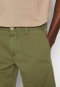 Manens överkropp i en beige skjorta instoppad i olivgröna byxor med handen i högra fickan, närbild på midja och höftområde.