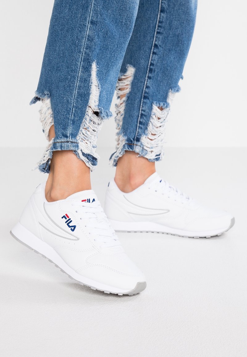 Baskets blanches FILA portées avec un jean bleu déchiré, montrant des ourlets effilochés et la cheville exposée sur un fond blanc uni.