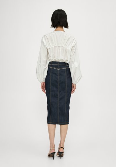 Blouse blanche avec des manches bouffantes assortie à une jupe crayon en denim foncé, ornée de coutures jaunes et d'une silhouette ajustée. Sandales à talons noires.