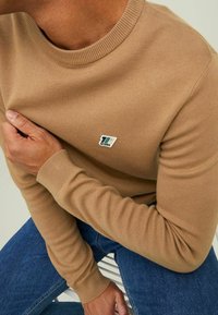 Vaaleanruskea neulottu villapaita, jossa on ribbireunainen pyöreä kaula-aukko ja pitkät hihat. Rinnassa pieni logopatchi. Yhdistetty sinisiin farkkuihin.