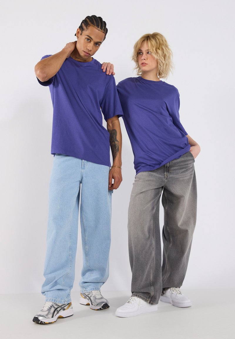 Violet kokvilnas t-krekli apvienoti ar gaiši ziliem un pelēkiem plašiem džinsiem. Apavi ir sudraba un baltas sporta kurpes ar melniem akcentiem.