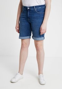 Denim-shortsit, keskiharmaansiniset, käännetyt lahkeenvarret, vetoketju ja napillinen kiinnitys, kaksi etutaskua, yhdistetty valkoisiin lenkkareihin ja yksiväriseen paitaan.
