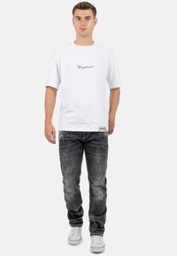 T-shirt bianca a maniche corte con logo in corsivo sul petto, abbinata a jeans neri sbiaditi e sneaker bianche con dettagli rossi.