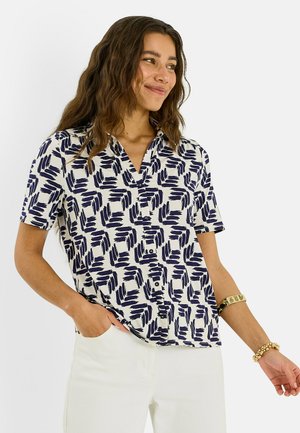 Mujer con cabello largo y ondulado que lleva una camisa blanca de manga corta con patrón abstracto negro, pantalones blancos y pulseras doradas, posando con la mano en el bolsillo.