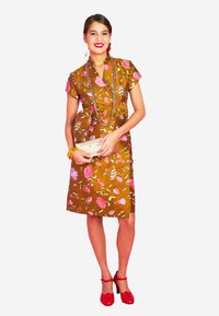 Braunes Blumenkleid mit pinken Blumen, kurzen Ärmeln und figurbetonter Passform. Rote Schuhe und eine metallische Clutch ergänzen das Outfit.