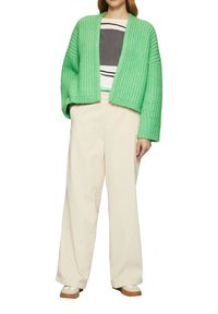 Grüner gestrickter Cardigan, offen vorne, strukturierter Stoff. Darunter ein gestreiftes grau-weißes Shirt. Cremefarbene weite Hose, braune Schuhe.