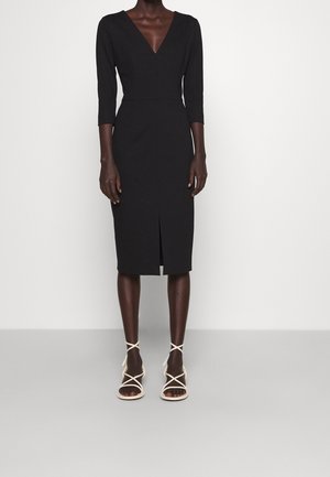 Femme portant une robe noire ajustée longueur genou avec des manches trois-quarts et une fente sur le devant, assortie à des sandales blanches à talons hauts et à brides.