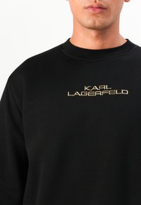 Sweatshirt preto com gola redonda canelada e logótipo "KARL LAGERFELD" bordado em dourado no peito. Textura de tecido suave.