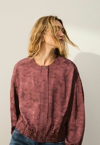 Chaqueta color burdeos con un patrón de hojas en relieve, cuello redondo, cierre de botones y cintura elástica; tela ligera con una textura suave.
