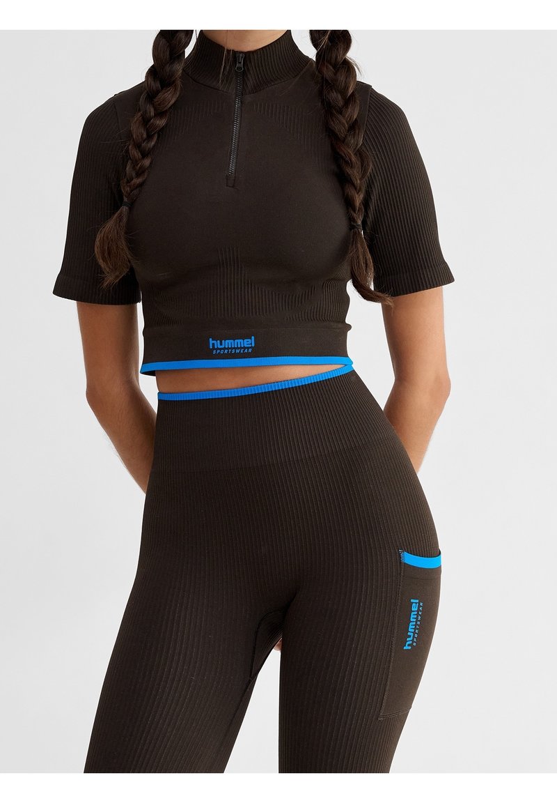 Personne avec deux tresses porte un ensemble de sport noir cintré Hummel avec des liserés bleus, composé d’un crop top à manches courtes et d’un legging taille haute.