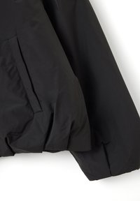 Schwarze gepolsterte Jacke mit schlankem Design, strukturiertem Stoff und einer seitlichen Tasche mit verstecktem Reißverschluss. Die Ärmel sind voluminös und weich strukturiert.