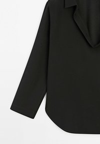 Manche longue noire de chemise et détail d'ourlet courbé sur un fond blanc uni.