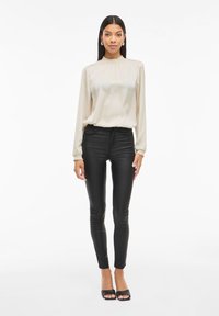 Blusa larga de manga larga en beige claro con un escote fruncido, combinada con pantalones negros ajustados y tacones abiertos negros. Textura suave.