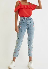 Czerwona bluzka typu off-shoulder z falbanami, zestawiona z jasnoniebieskimi spodniami z wysokim stanem, zdobionymi kolorowym haftem kwiatowym, oraz białymi trampkami.