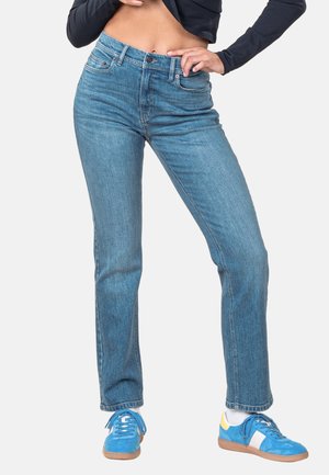 Frau trägt blaue Jeans mit mittlerer Taille und blaue Sneakers, steht mit einer Hand in der Hüfte und einem schwarzen, bauchfreien Oberteil.