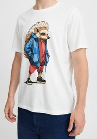 Hvid bomuld t-shirt med et pindsvin-tryk i blå jakke, rød skjorte og sorte sko. Korte ærmer og rund halsudskæring.
