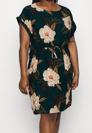 Mujer con vestido verde oscuro hasta la rodilla con un gran estampado floral blanco y cintura con cordón, de pie frente a un fondo blanco liso.