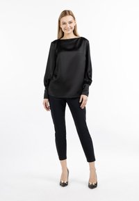 Schwarze Satinbluse mit langen Ärmeln, lockerer Passform und rundem Halsausschnitt, kombiniert mit schmal geschnittenen schwarzen Hosen und glänzenden schwarzen High Heels.