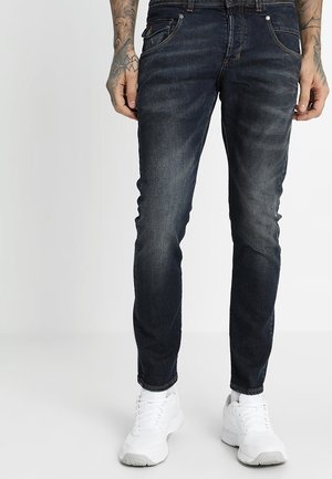 Homme portant un jean slim bleu foncé et des baskets blanches, debout contre un fond clair uni avec les mains tatouées visibles.