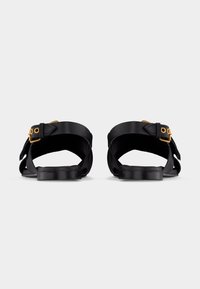 Μαύρα σανδάλια με χρυσές αγκράφες, φωτογραφημένα από πίσω σε λευκό φόντο, με μαξιλαρωτές επίπεδες σόλες.