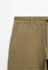 Zone de la taille d’un pantalon kaki avec poche avant en biais, passants de ceinture, coutures visibles et fermeture par bouton noir sur fond blanc.