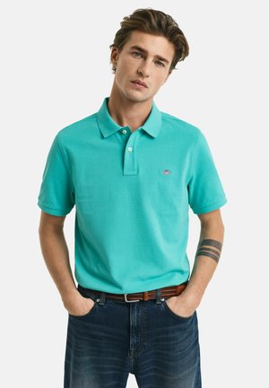 Turkis polo skjorte med korte ærmer, knaplukning og broderet logo. Sammen med mørk denim jeans og et brunt bælte.