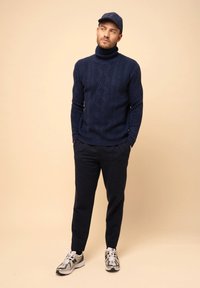 Navyfarbener Strickpullover mit Rollkragen und strukturiertem Muster, kombiniert mit dunklen Hosen und gemusterten Sneakers. Die Mütze ist navyfarben mit einem flachen Schirm.