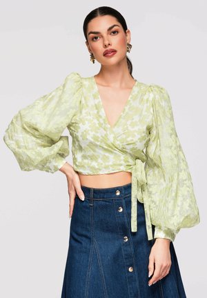 Femme portant un chemisier portefeuille léger vert clair à fleurs avec des manches bouffantes et une jupe en denim bleu foncé taille haute avec boutons à l'avant.