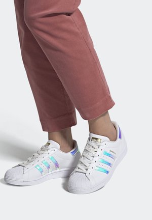 Osoba nosící bílé tenisky Adidas Superstar s duhově modro-fialovými pruhy a fialovými detaily, doplněné o růžové kalhoty s krátkou délkou.