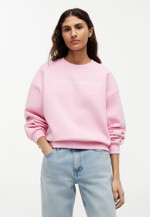 Giovane donna che indossa una felpa rosa chiaro Tommy Hilfiger con logo e jeans azzurri, in piedi con una mano in tasca.