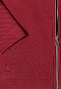 Close-up of a red textured fabric jacket with a silver zipper and a snap-button pocket flap.