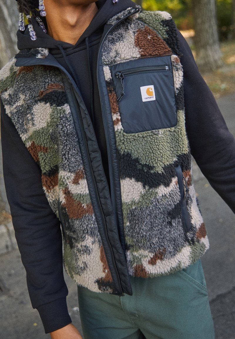 Chaleco de forro polar con patrón de camuflaje multicolor en verde, marrón y gris. Presenta un bolsillo frontal con cremallera negra contrastante y cremallera delantera.