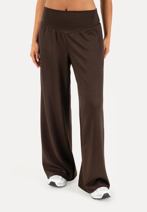 Donna con pantaloni larghi marrone scuro e sneakers bianche, in piedi davanti a uno sfondo chiaro e semplice.