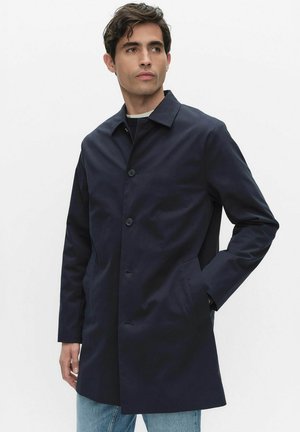 Man die een donkerblauwe enkelgeknopte jas draagt, staand met één hand in de zak, kijkend naar de zijkant tegen een effen achtergrond.