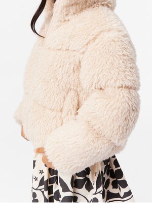 Kobieta ubrana w jasnobeżową, puszystą kurtkę na czarno-białą spódnicę w kwiatowy wzór, stojąca na tle białym.