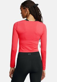 Nainen, jolla on letitetyt hiukset, pukeutuneena punaiseen pitkähihaiseen crop-toppiin ja mustiin Under Armour -legginsseihin, nähtynä takaa valkoista taustaa vasten.