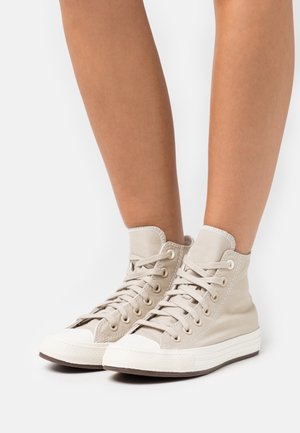 Person wearing beige high-top canvas sneakers with white rubber soles and toe caps, standing on a white surface.