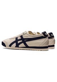 Beżowe skórzane sneakersy z czarnymi paskami i czarnymi wstawkami na pięcie, widok z boku pokazujący niskoprofilowy design i teksturowane gumowe podeszwy.