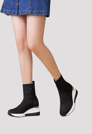 Bottines noires en tricot avec un talon en coin, présentant une semelle blanche et un accent métallique sur le côté. La texture est côtelée et ajustée.