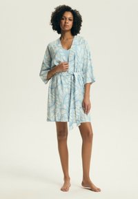 Žena kučeravých vlasov, ktorá nosí svetlomodrý kvetovaný župan zviazaný v páse, stojaca bosá pred jednoduchým pozadím.